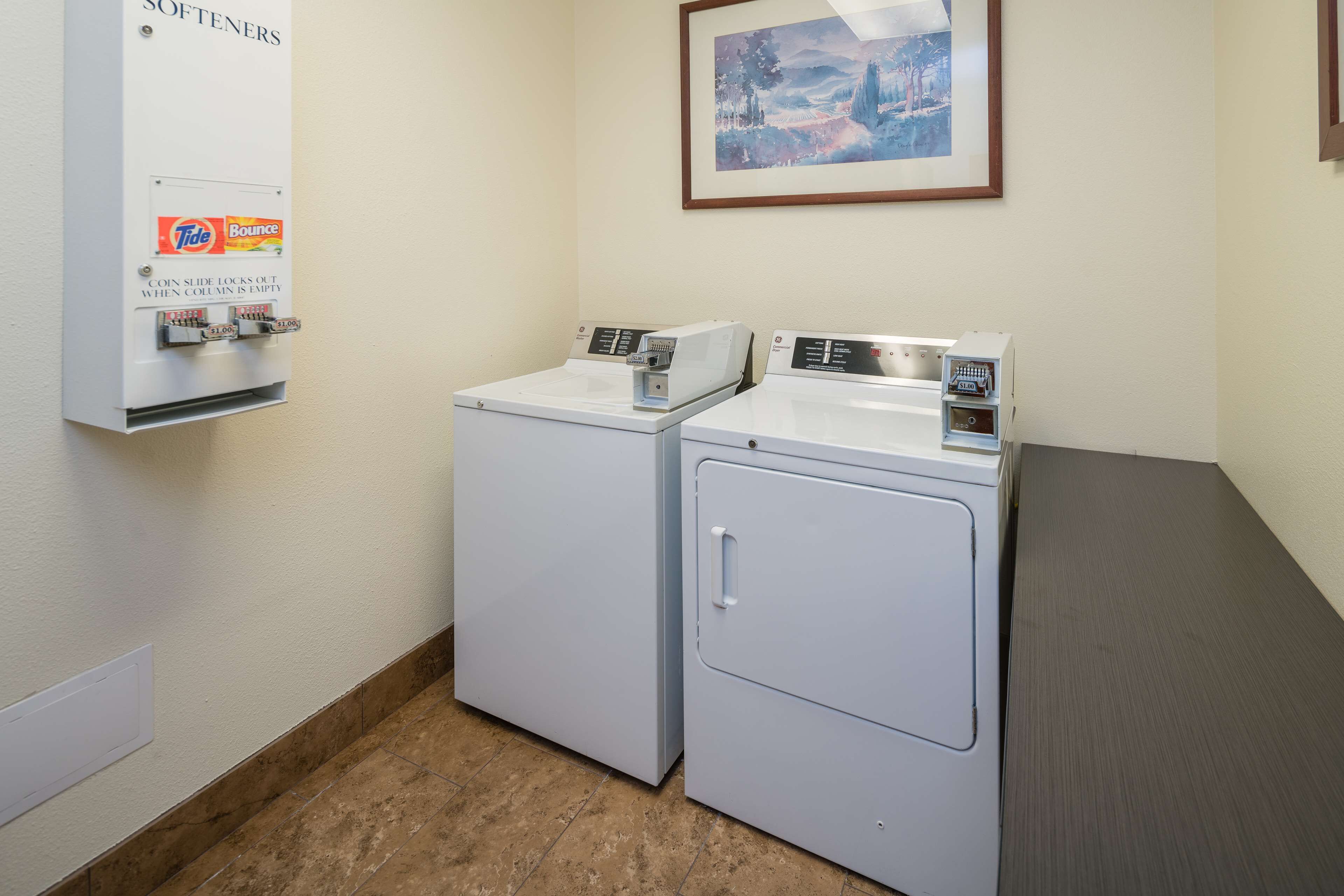 A laundry area with two front-loading washers and dryers, a coin-operated softener dispenser, and a framed landscape painting on the wall.