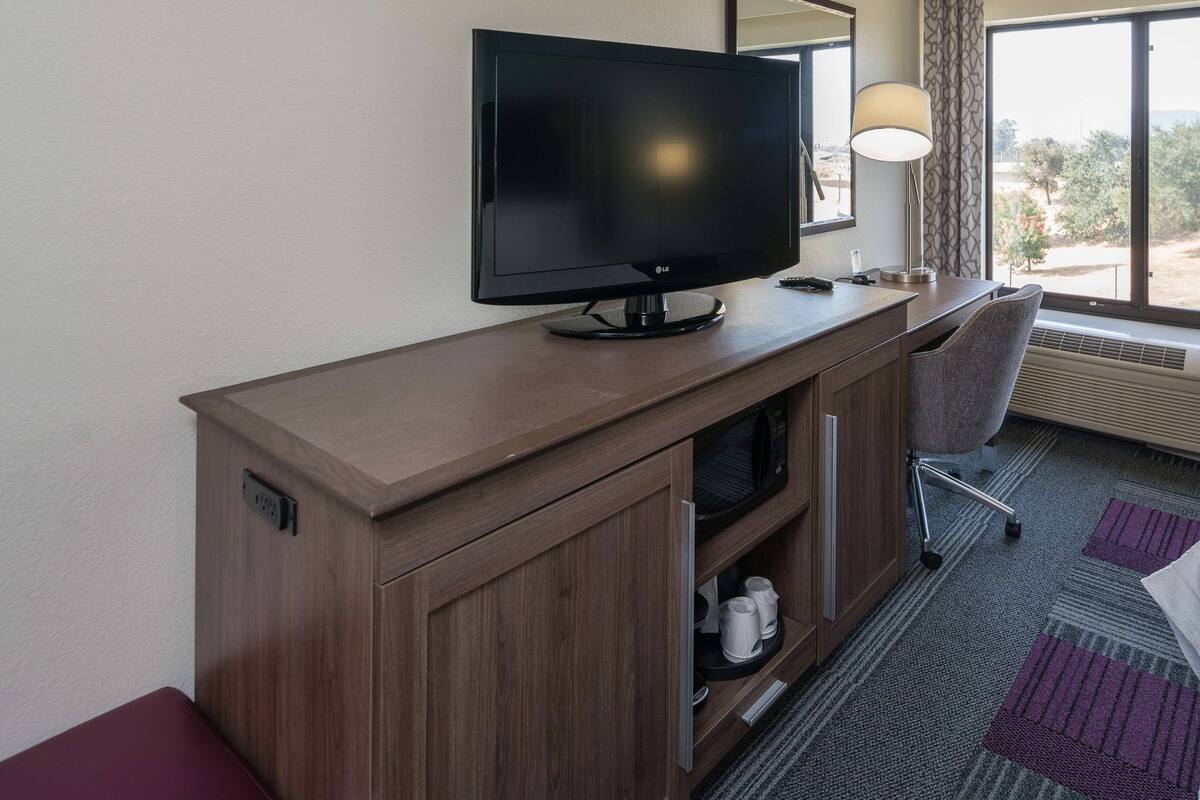 A hotel room with a wooden desk, a flat-screen TV, a chair, a microwave, and a window with a view of trees.