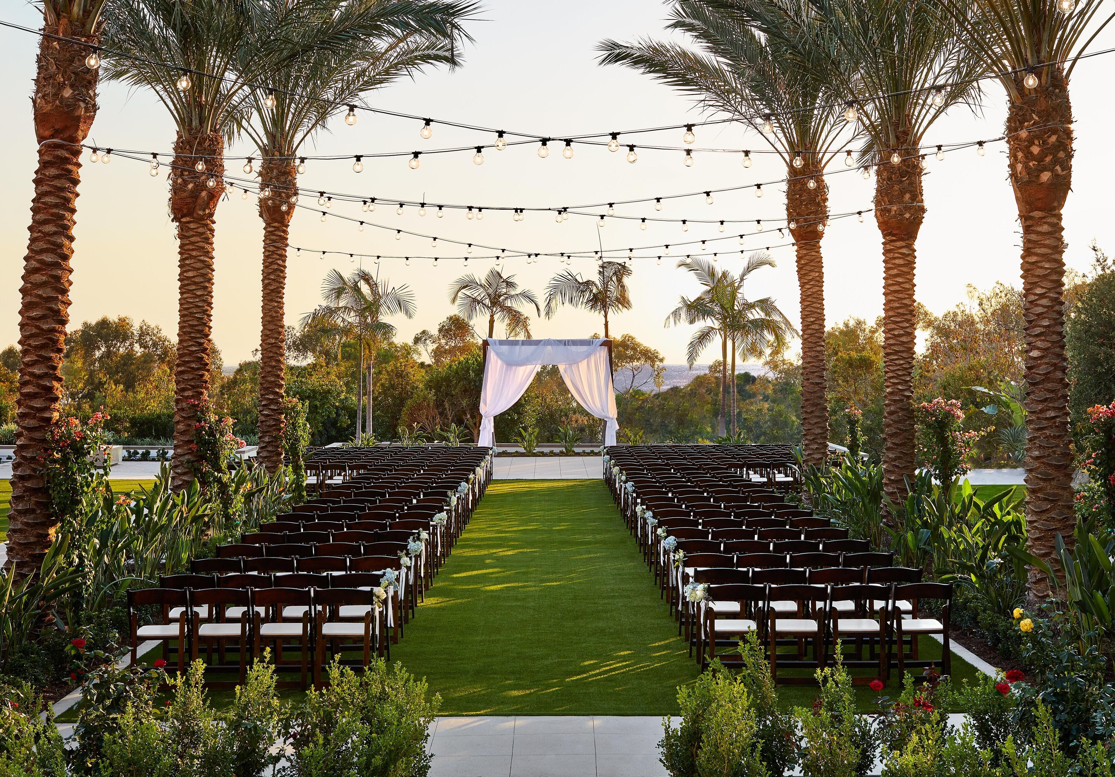 An outdoor wedding venue with rows of wooden chairs and a green carpet aisle, surrounded by palm trees and string lights.