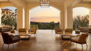 Un balcón amplio con vista a un paisaje urbano al atardecer, amueblado con sillas de mimbre y una mesa.