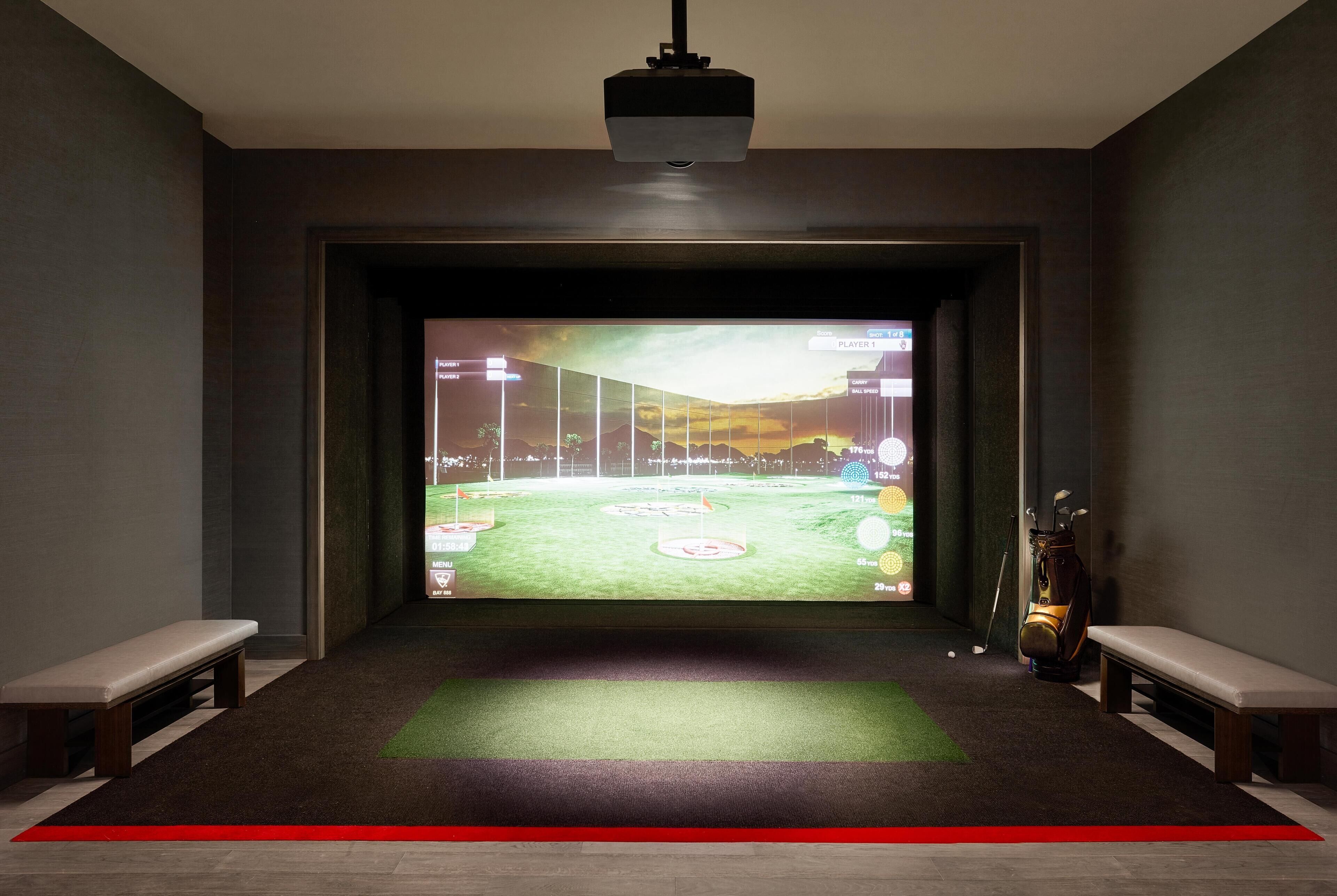 A room with a large screen displaying a golf game, a golf bag, and two benches.
