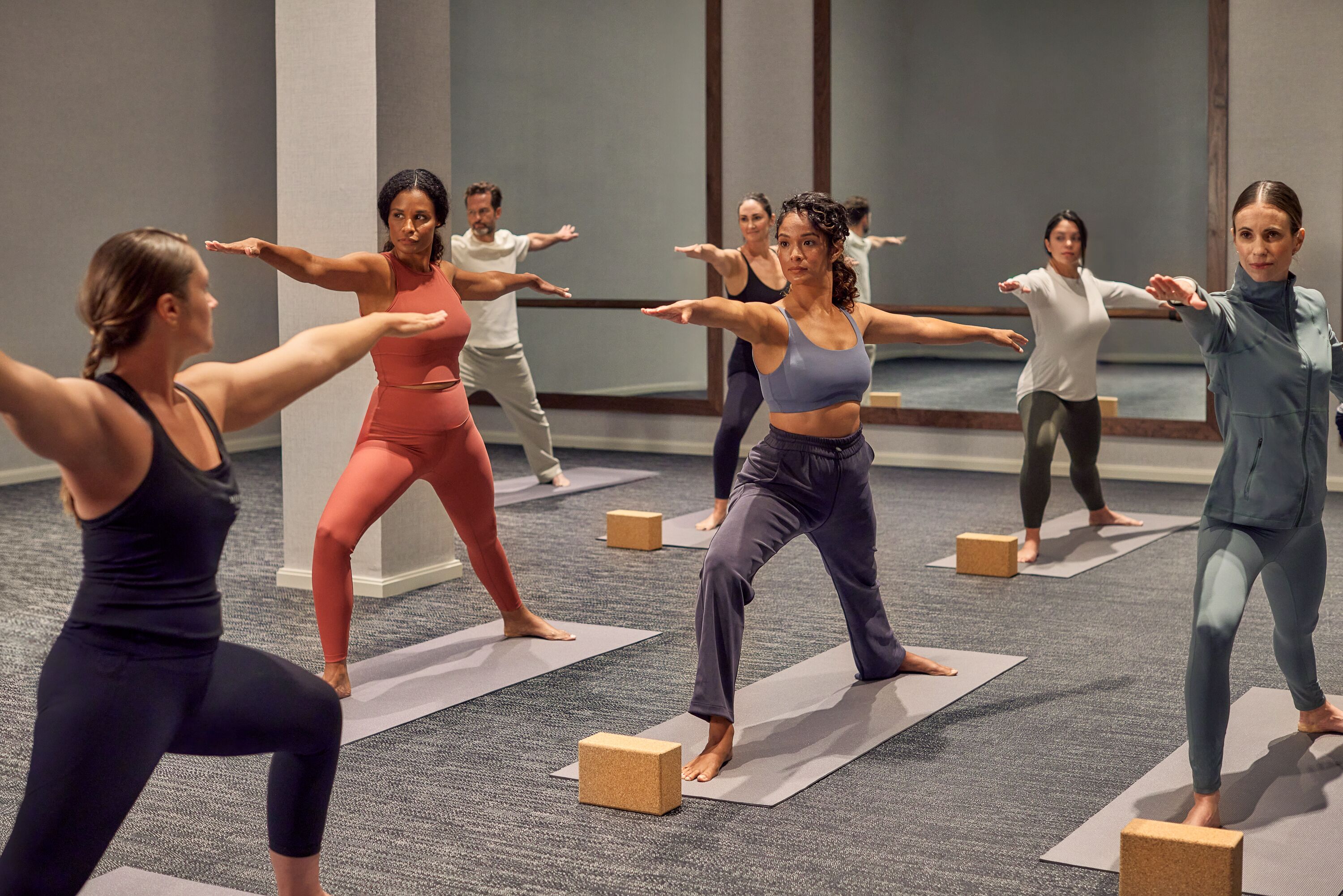 A group of people in a room with carpeted floors, standing on yoga mats with wooden blocks, performing a yoga pose.