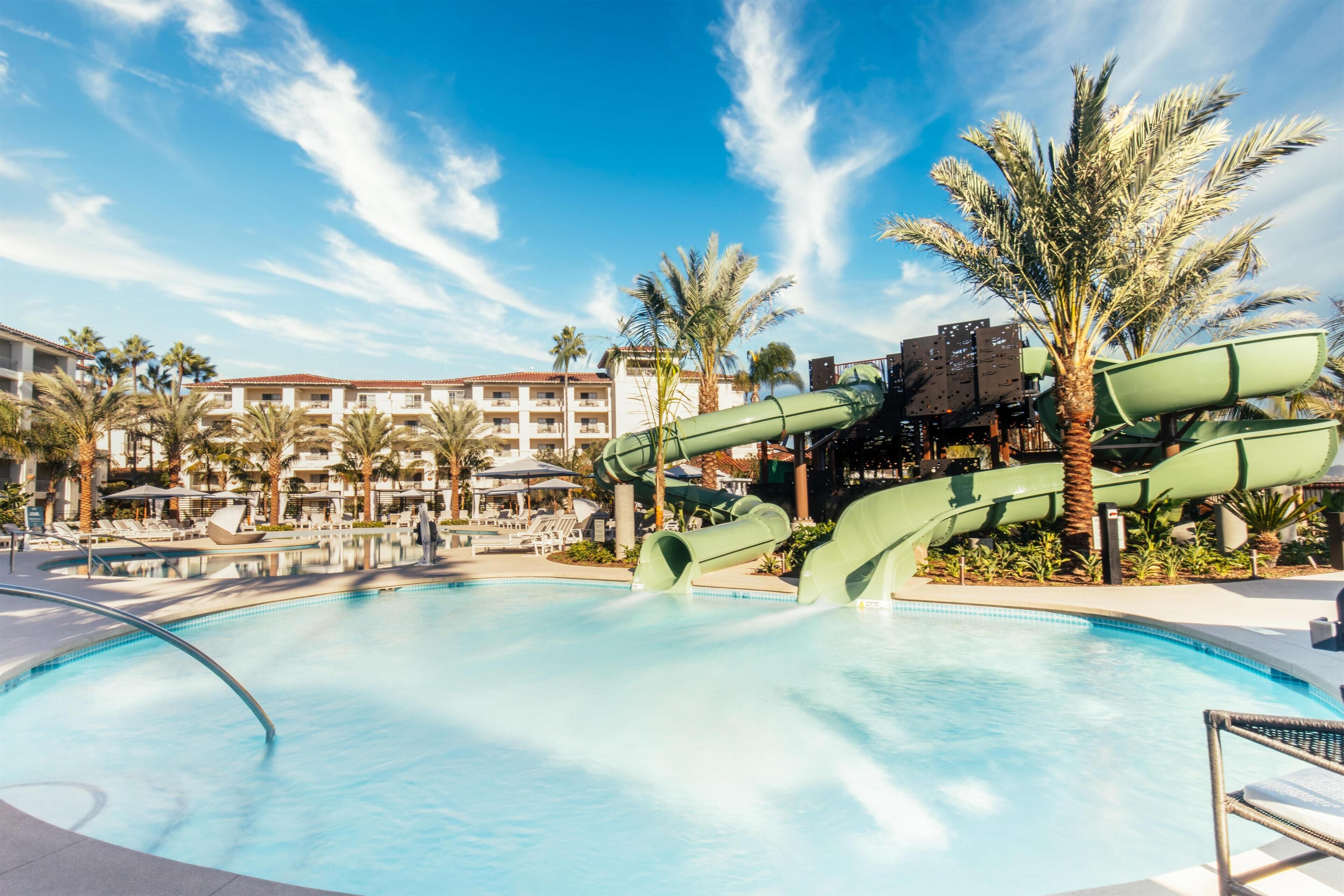 A swimming pool with a water slide, palm trees, and a building with balconies in the background.