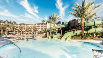 A swimming pool with a water slide, palm trees, and a building with balconies in the background.