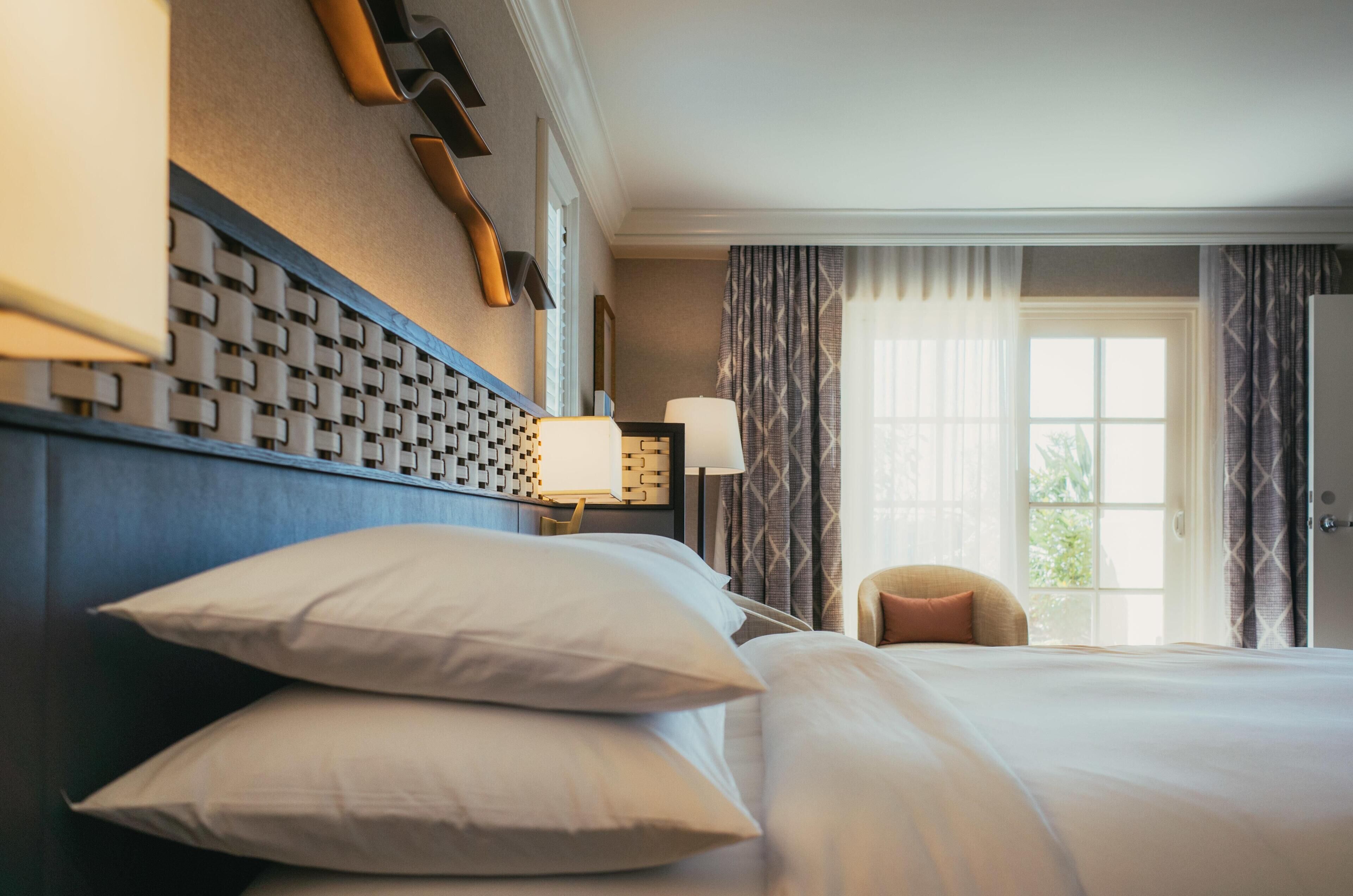 Ein Hotelzimmer mit einem großen Bett, einem Nachttisch mit Lampe, einem Sessel und einem Fenster mit Vorhängen.