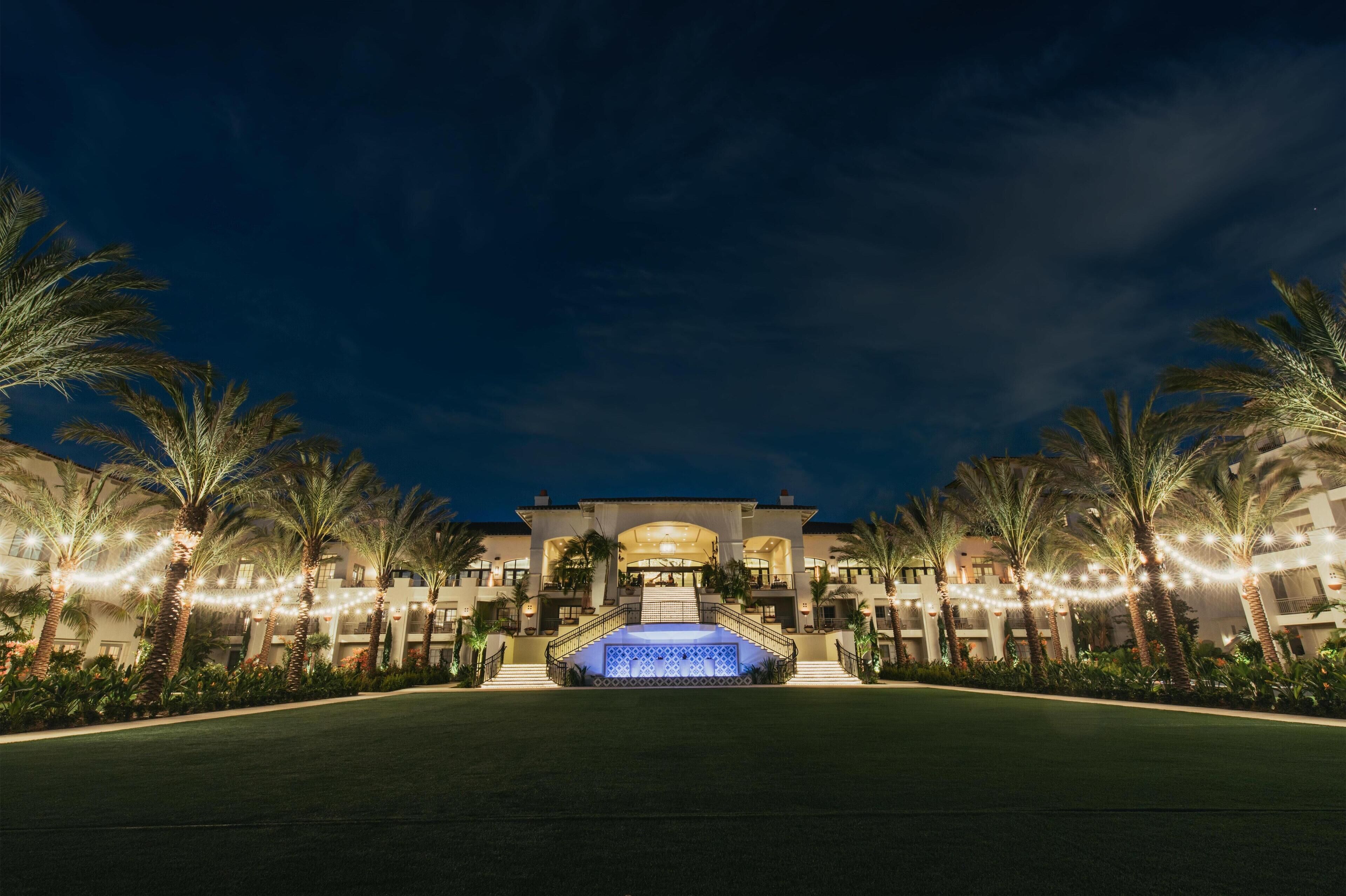 A large, well-lit hotel with a central fountain, surrounded by palm trees and a manicured lawn.