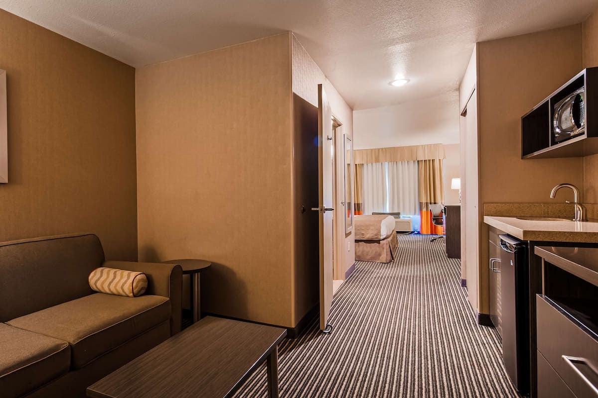 A hotel room with a sofa, a small table, a kitchenette, and a bed with curtains.