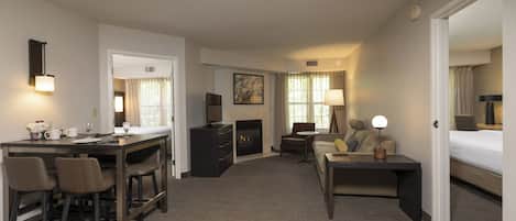 A hotel room with a dining area, a sofa, a small table with a lamp, and a fireplace.