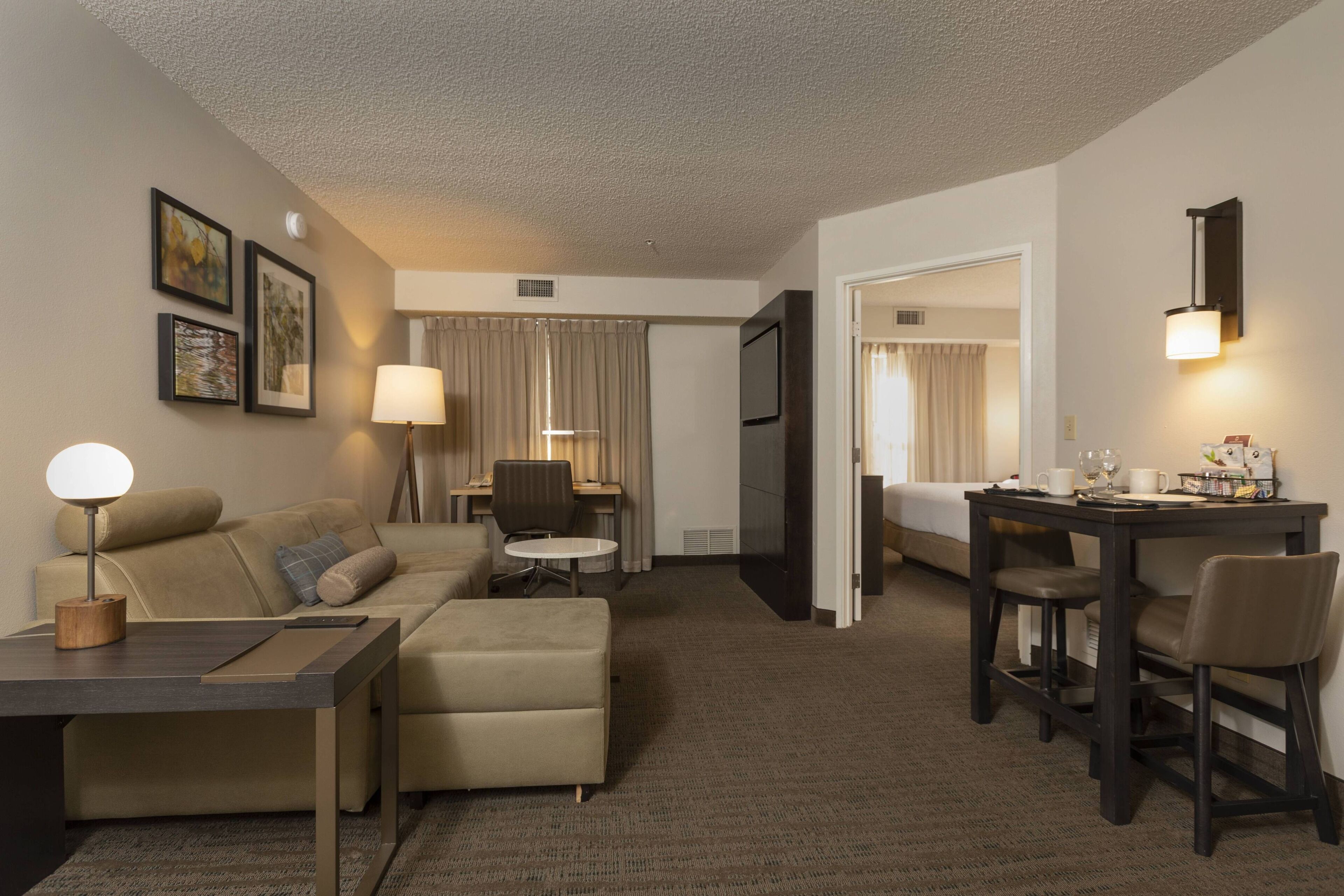 A hotel room with a sofa, a small table, a desk with chairs, a TV, and a lamp.