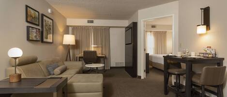 A hotel room with a sofa, a small table, a desk with chairs, a TV, and a lamp.