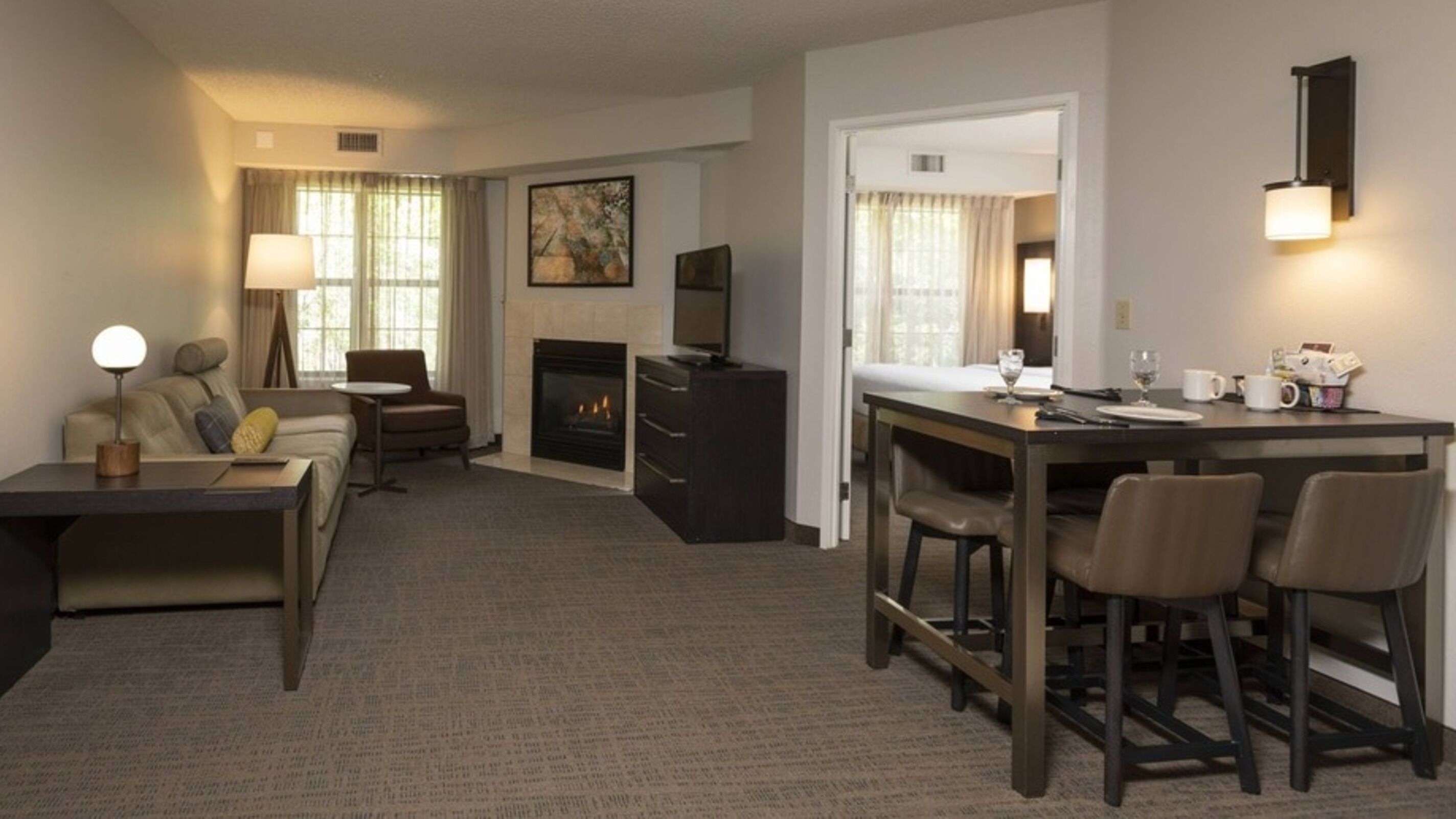 A hotel room with a sofa, a dining table with chairs, a fireplace, a TV, and a wall-mounted light fixture.