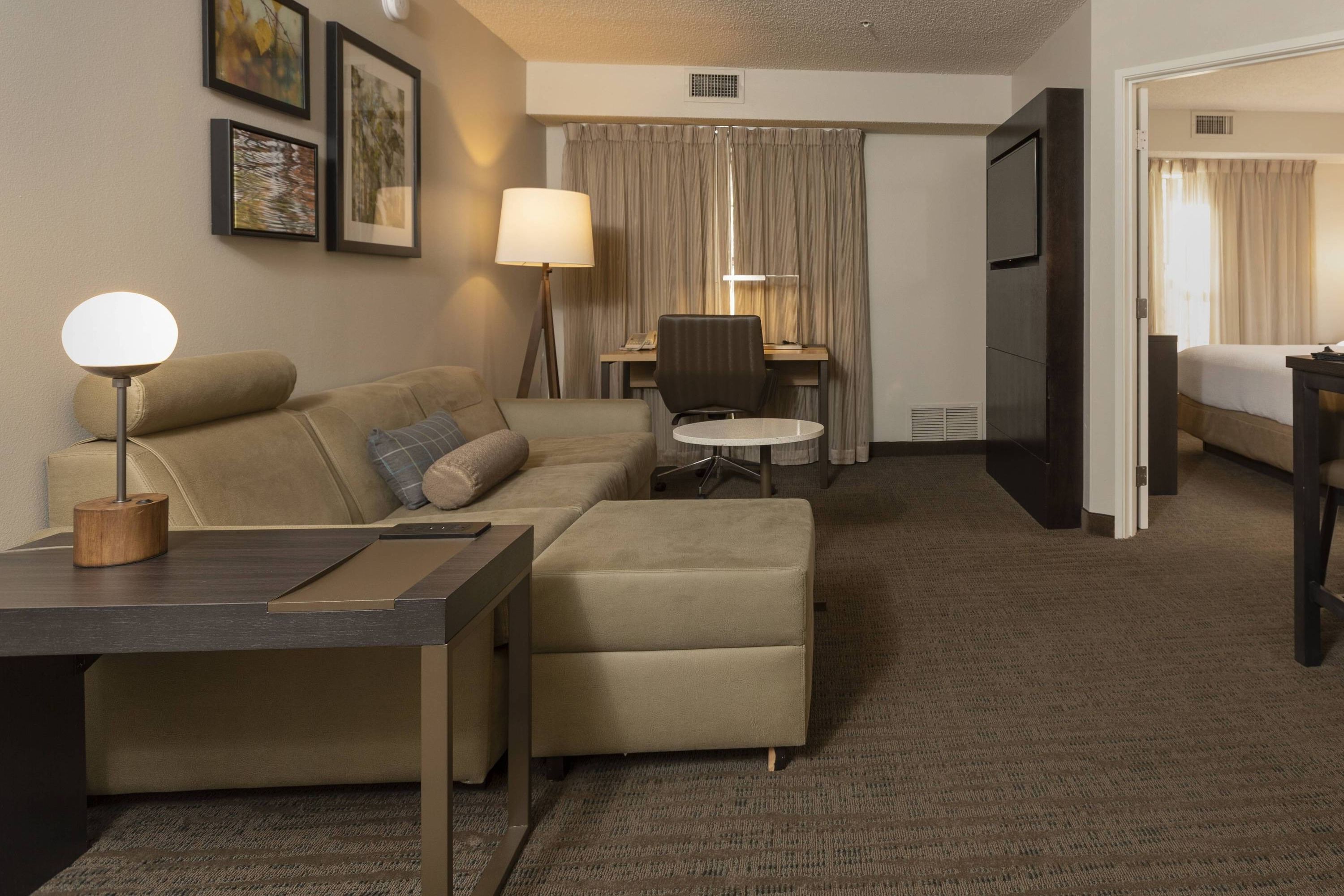 Een hotelkamer met een bank, poef, bureau, lamp, stoel en een bed op de achtergrond.