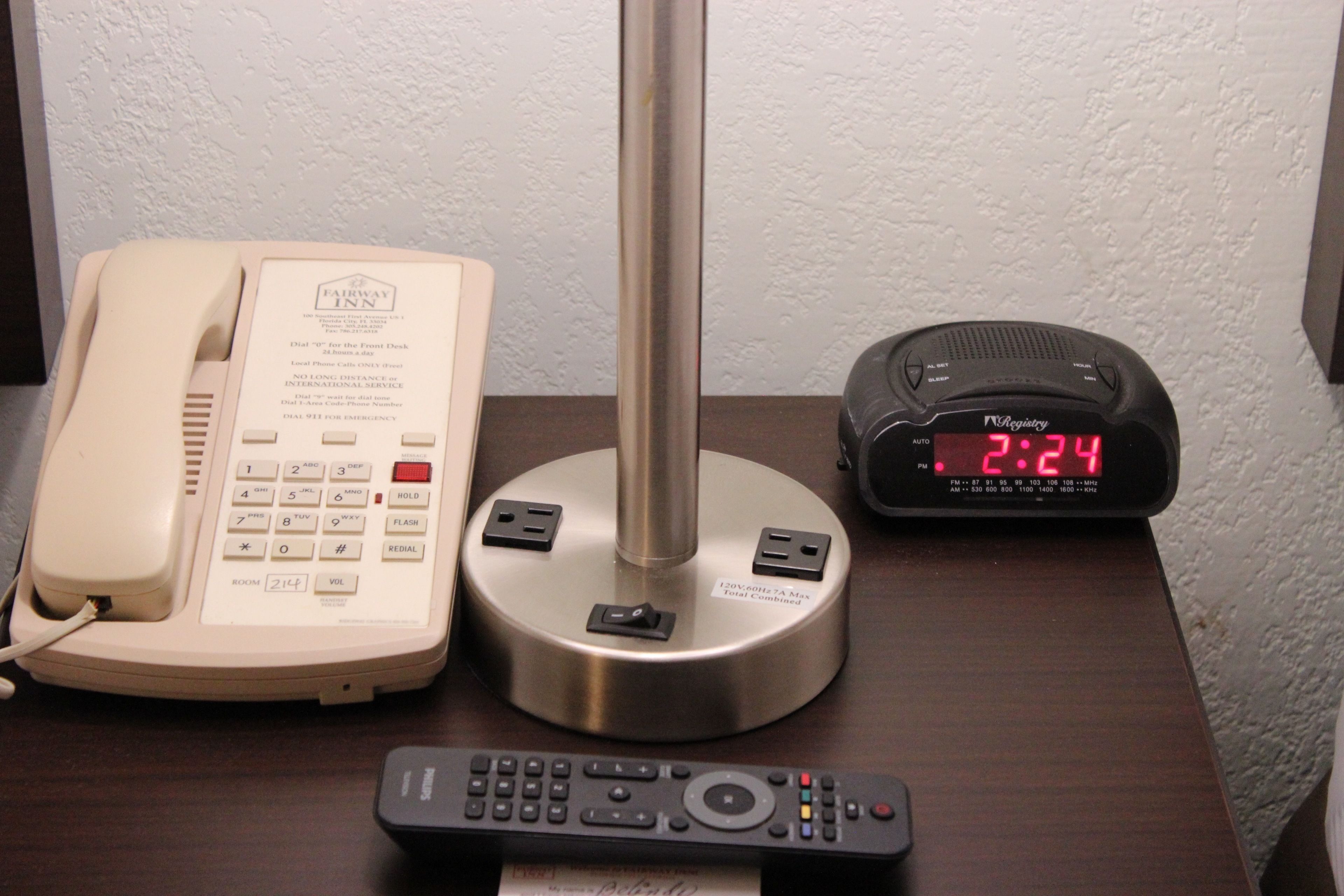 A hotel room with a bedside telephone, a digital clock, and a remote control on a wooden nightstand.