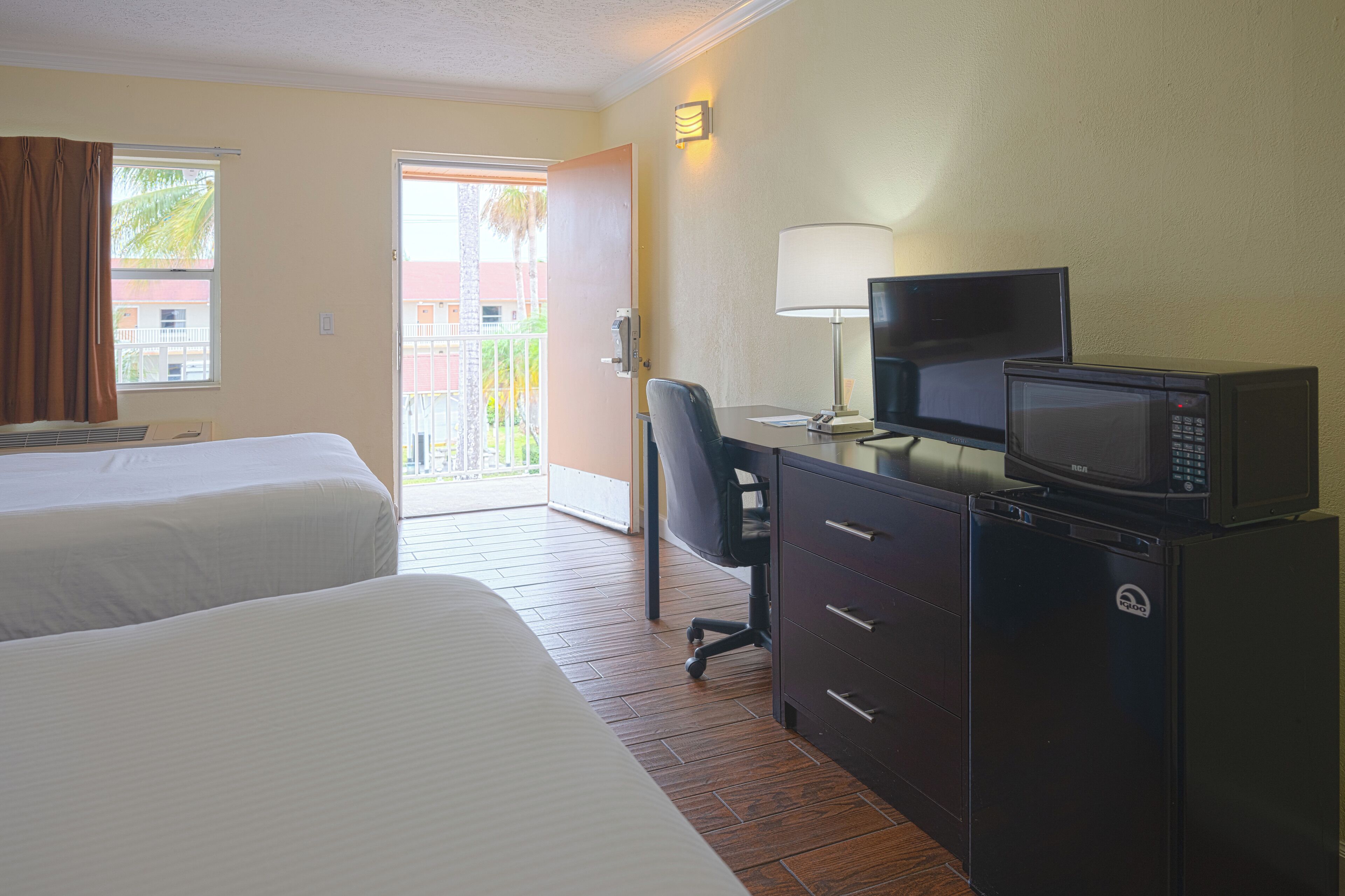 Ein Hotelzimmer mit zwei Betten, einem Schreibtisch mit Computer und Mikrowelle sowie Blick auf ein Gebäude und Palmen durch die Tür.