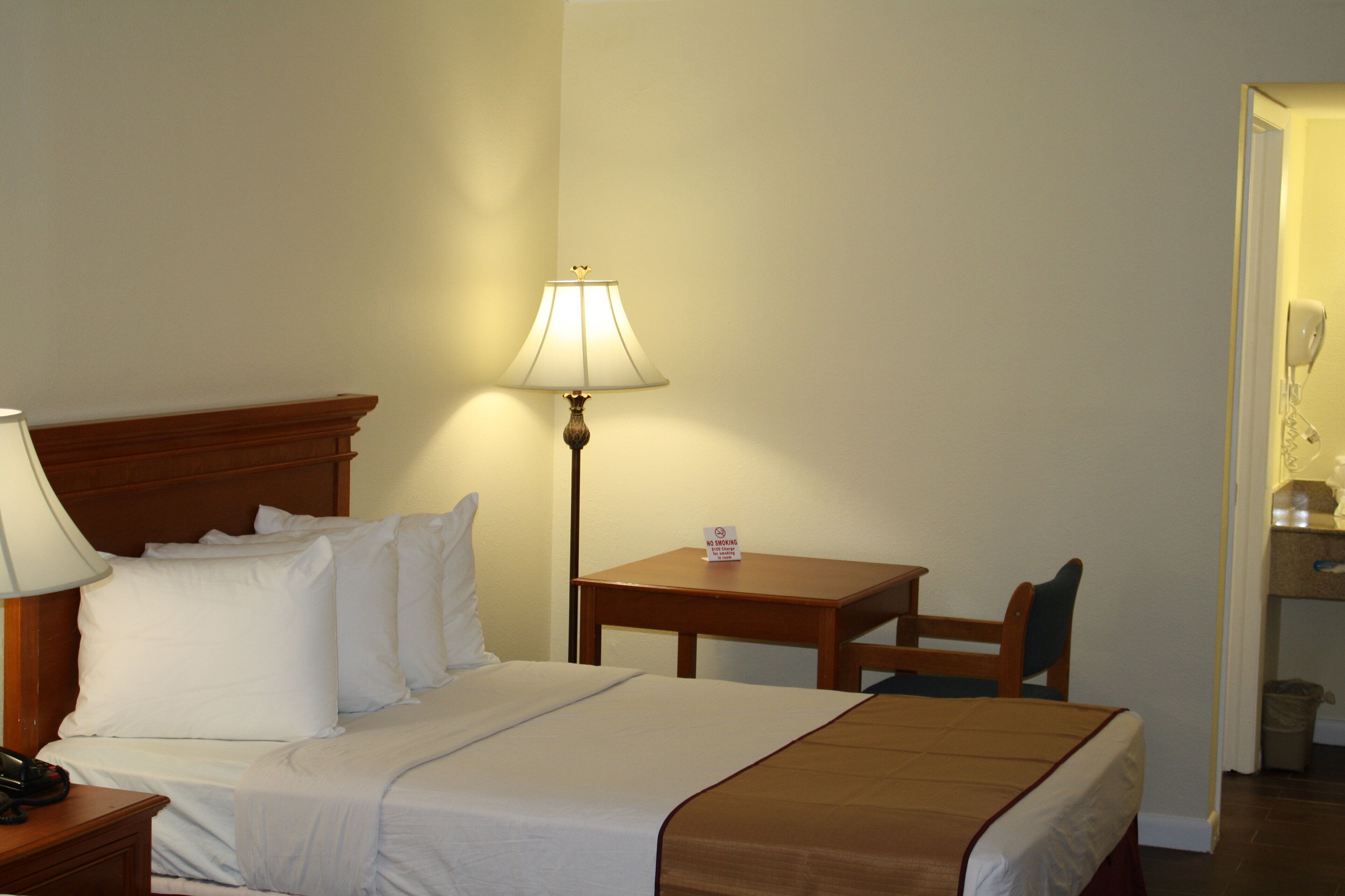 A hotel room with a bed, bedside table, lamp, chair, and a small table with a sign.
