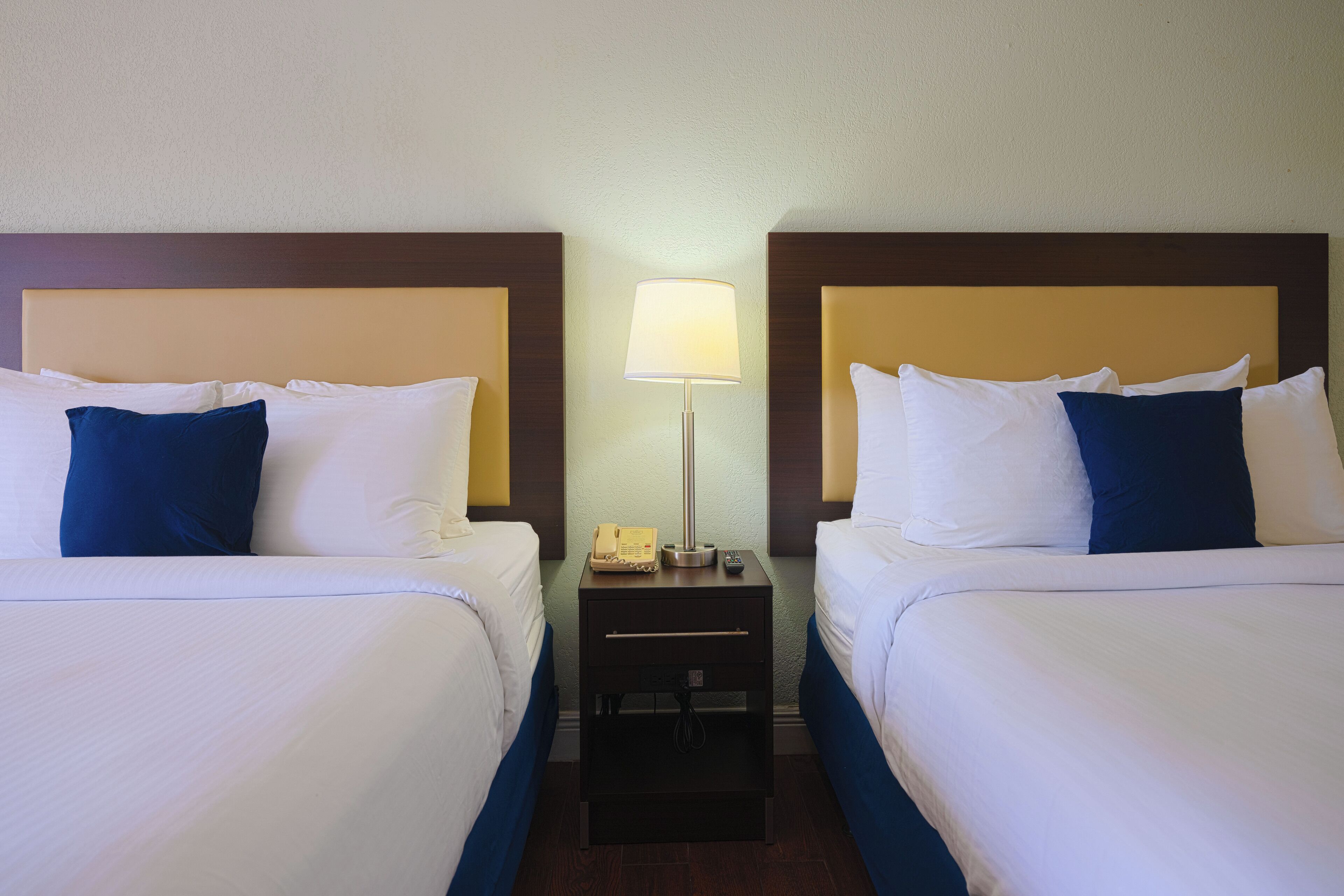 Quarto de hotel com duas camas, cada uma com roupa de cama branca e azul, uma mesa de cabeceira com abajur e telefone.