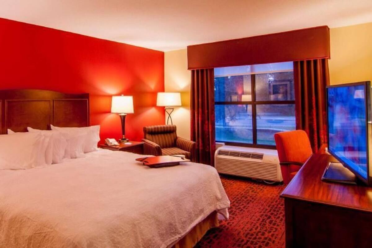 A hotel room with a large bed, two bedside tables, a desk with a chair, a window with curtains, and an air conditioning unit.
