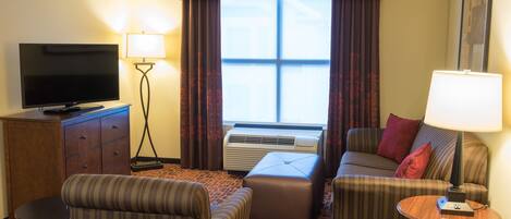 A hotel room with a television, a sofa, an ottoman, a lamp, and a window with curtains.