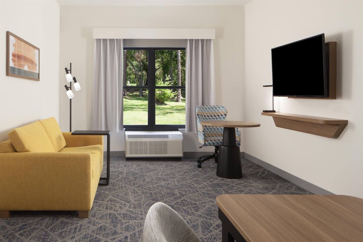 A hotel room with a yellow sofa, a small table, a TV mounted on the wall, and a window with curtains.