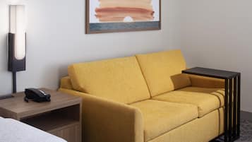 Habitación de hotel con sofá amarillo, una mesita de noche con lámpara, una obra de arte enmarcada en la pared y una cama parcialmente visible.