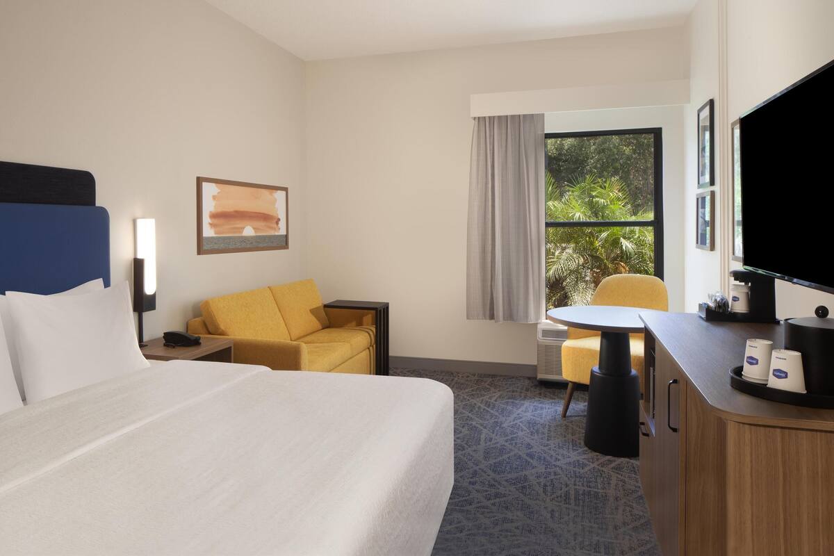 A hotel room with a bed, a desk with a chair, a TV, and a window with a view of greenery.