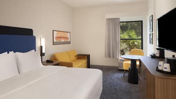 A hotel room with a bed, a desk with a chair, a TV, and a window with a view of greenery.