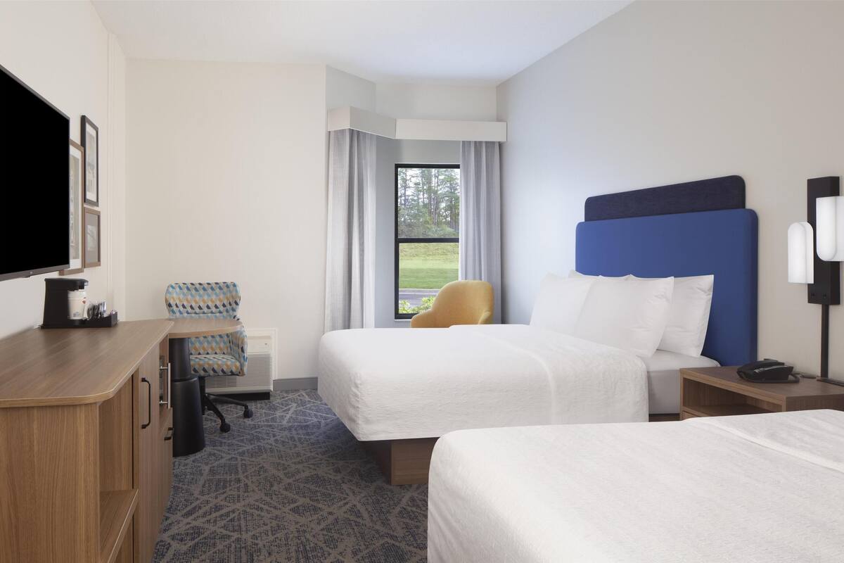 A hotel room with two beds, a desk, a chair, a TV, and a window with curtains.