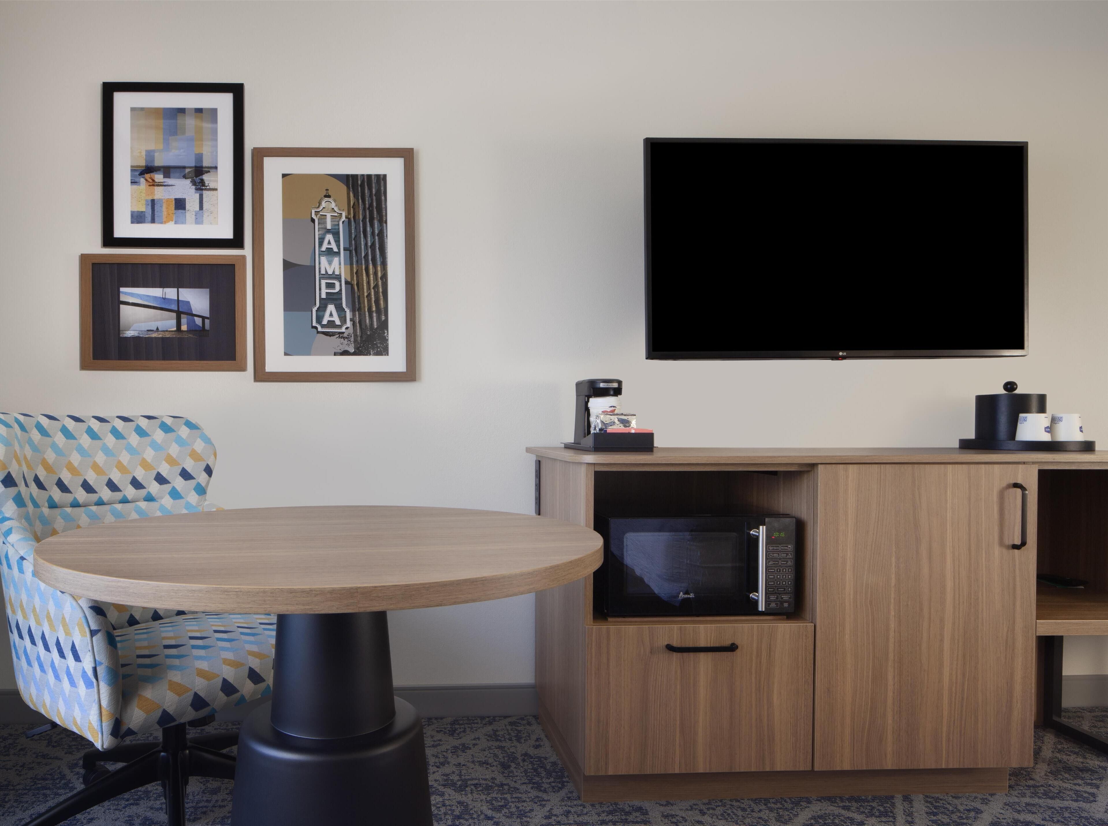 A hotel room with a round wooden table, a sofa with a geometric pattern, a flat-screen TV, a coffee maker, a microwave, and framed artwork on the wall.