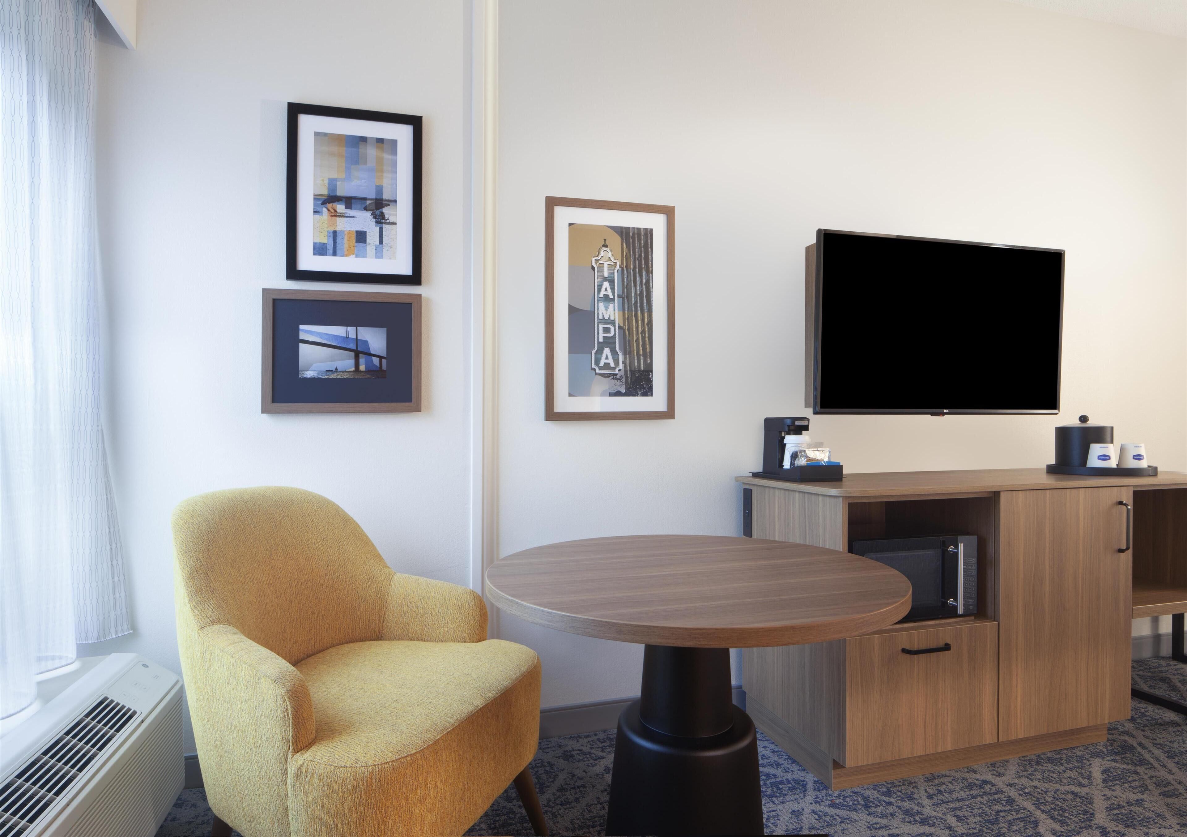 A hotel room with a yellow armchair, a round table, a flat-screen TV, framed pictures on the wall, and a minibar.