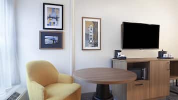 A hotel room with a yellow armchair, a round table, a flat-screen TV, framed pictures on the wall, and a minibar.