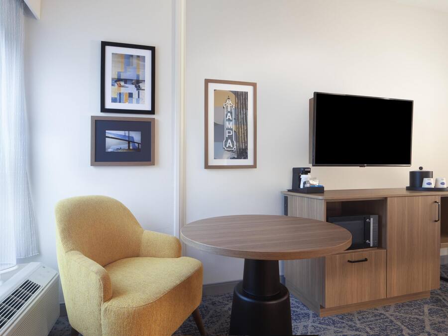A hotel room with a yellow armchair, a round table, a flat-screen TV, framed pictures on the wall, and a minibar.