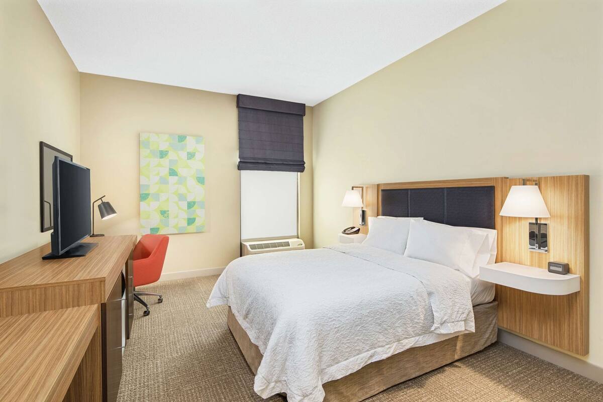 A hotel room with a bed, bedside table, lamp, desk with a computer, chair, and a window with a curtain.