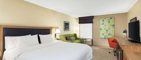 A hotel room with a large bed, a desk with a television, a green chair, and a painting on the wall.