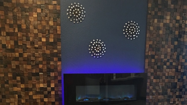 Ein Zimmer mit zwei roten Sesseln, einem kleinen runden Tisch, einer blauen Wand mit Holz-Mosaikstrukturen und einem an der Wand befestigten Flachbildfernseher.