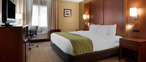 A hotel room with a large bed, a desk with a television, a chair, and a desk lamp.
