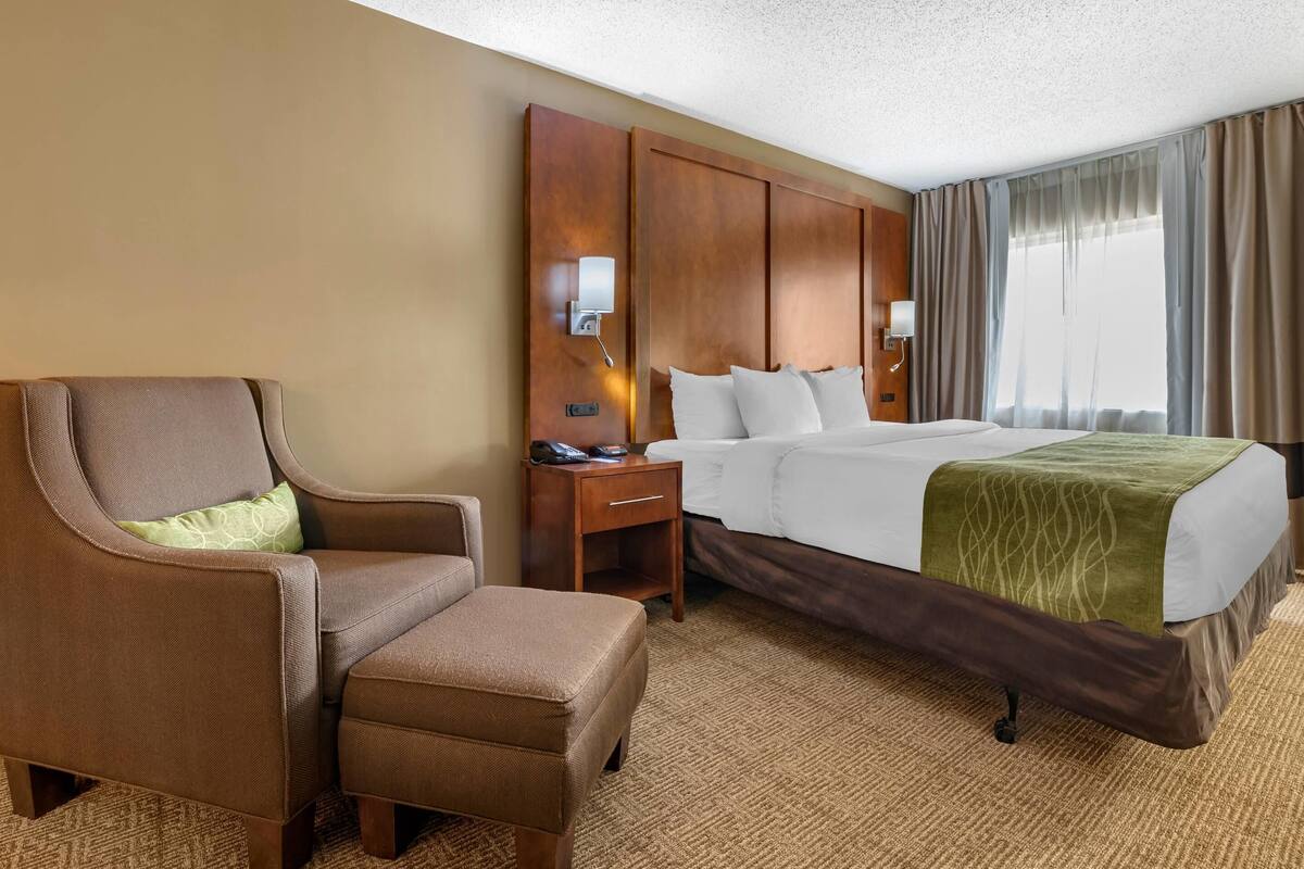 A hotel room with a bed, a brown armchair, a nightstand, and a large window with curtains.