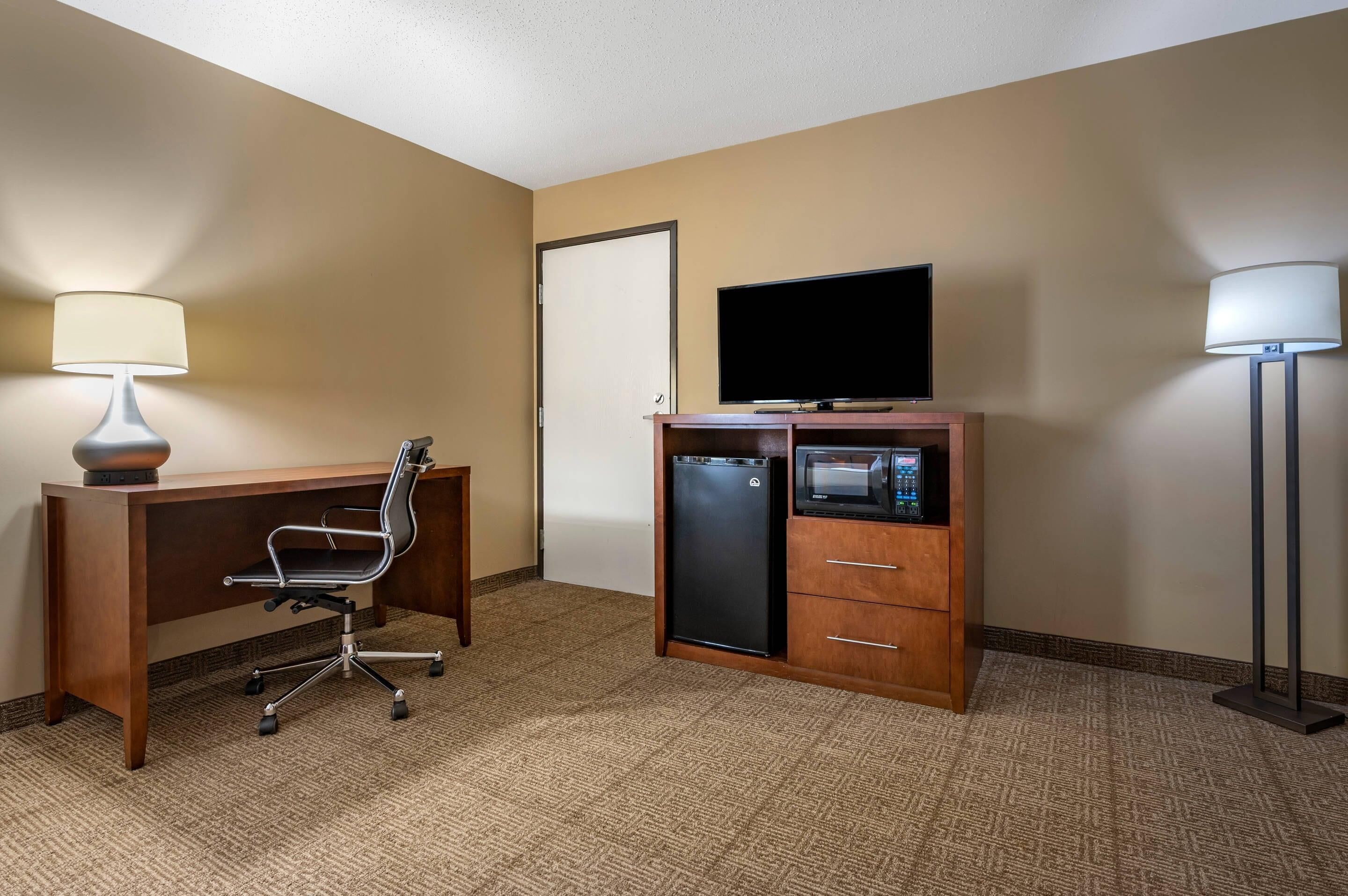 Une chambre d’hôtel équipée d’un bureau en bois, d’un fauteuil de bureau noir, d’un téléviseur à écran plat, d’un mini-réfrigérateur, d’un four à micro-ondes et d’une lampe sur pied.