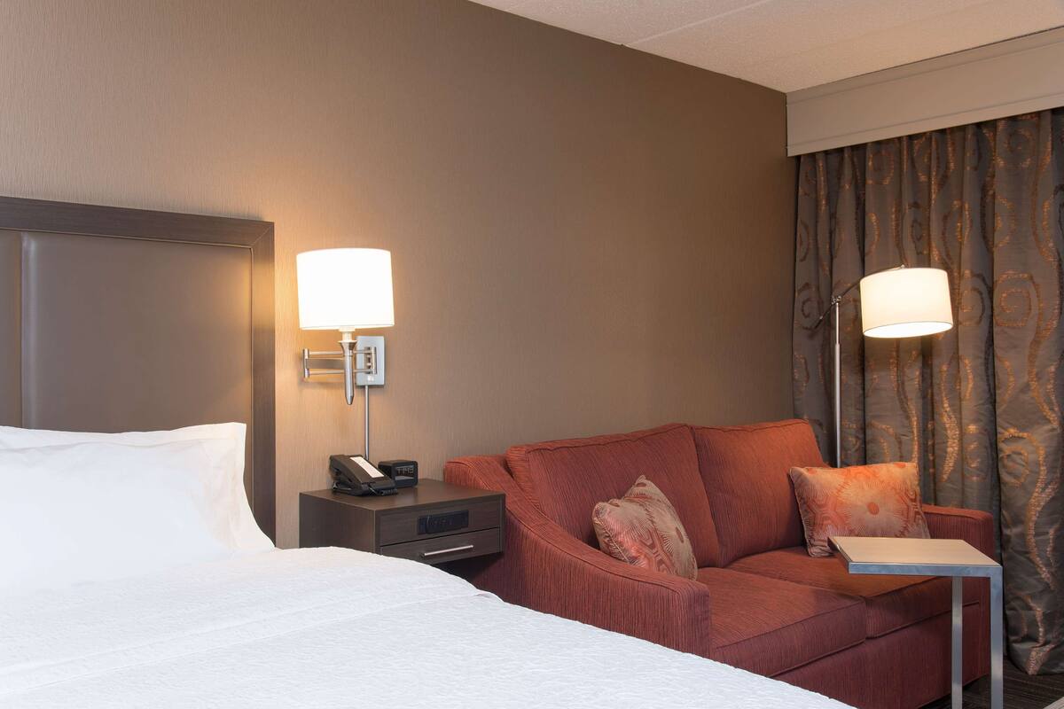 A hotel room with a bed, a sofa, a nightstand with a lamp, and a small table.