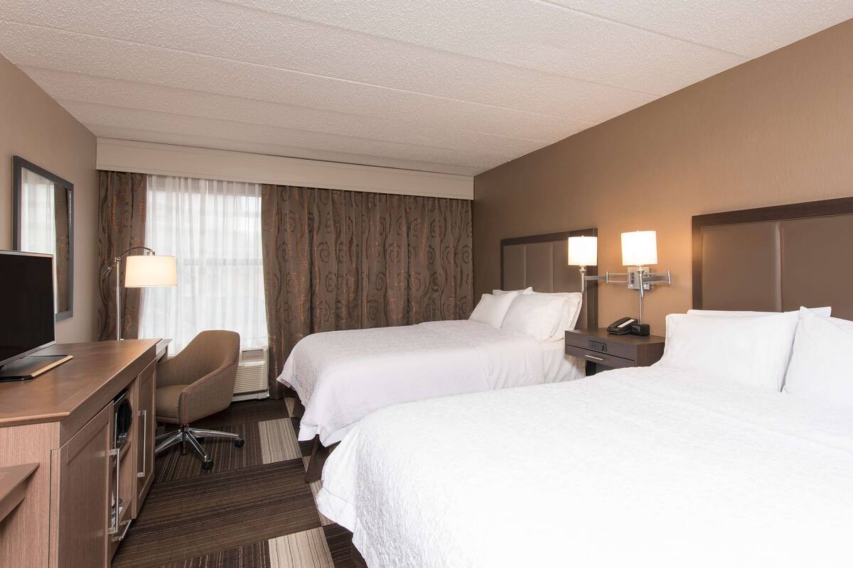 A hotel room with two beds, a desk, a chair, a TV, and a window with curtains.