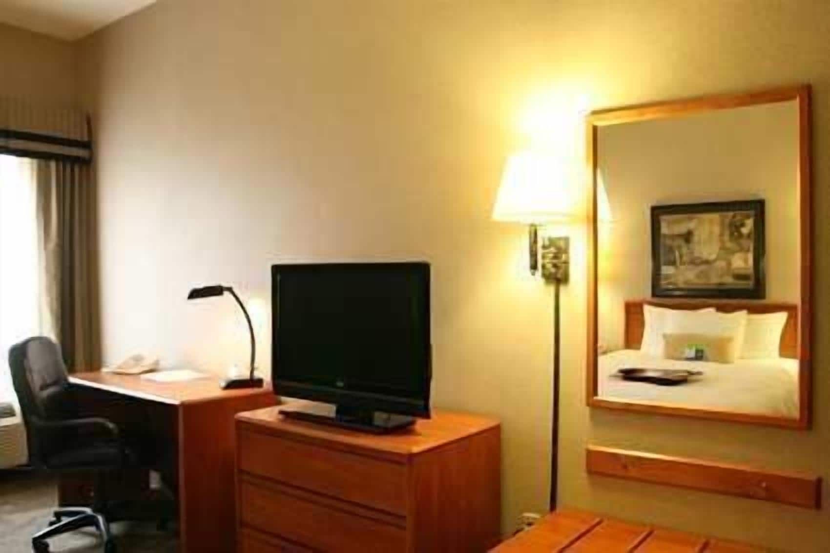 A hotel room with a desk, a flat-screen TV, a lamp, a bed with a headboard, and a mirror.