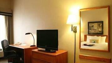 A hotel room with a desk, a flat-screen TV, a lamp, a bed with a headboard, and a mirror.