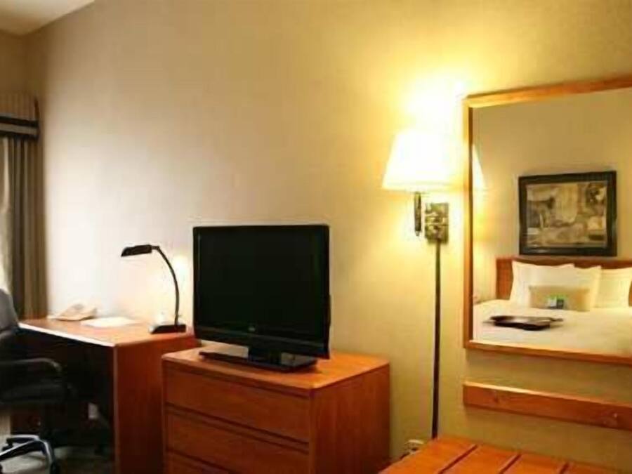 A hotel room with a desk, a flat-screen TV, a lamp, a bed with a headboard, and a mirror.