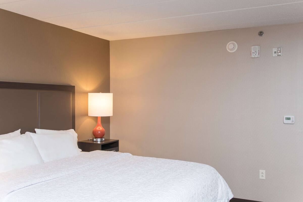 A neatly made bed with white linens, a red lamp on a bedside table, and a wall with a thermostat and a smoke detector.