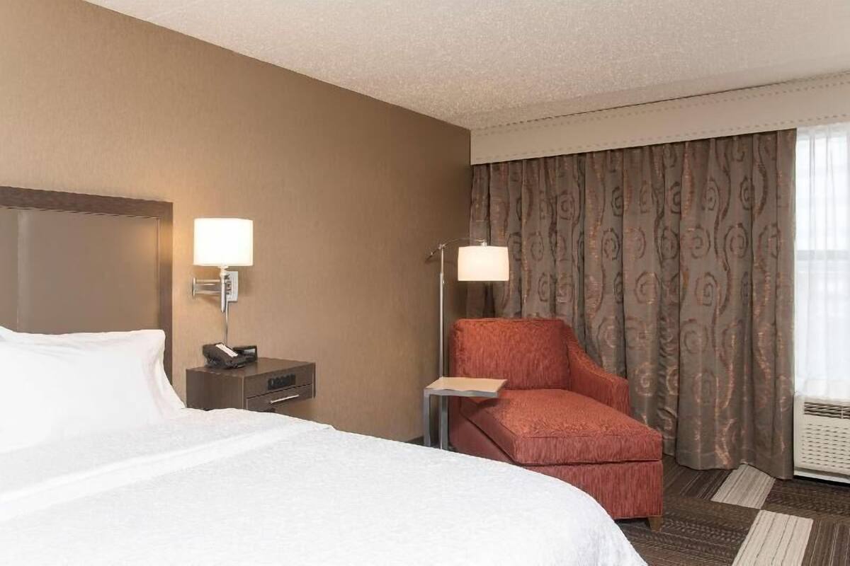 A hotel room with a bed, two bedside lamps, a chair, a nightstand, and a window with curtains.