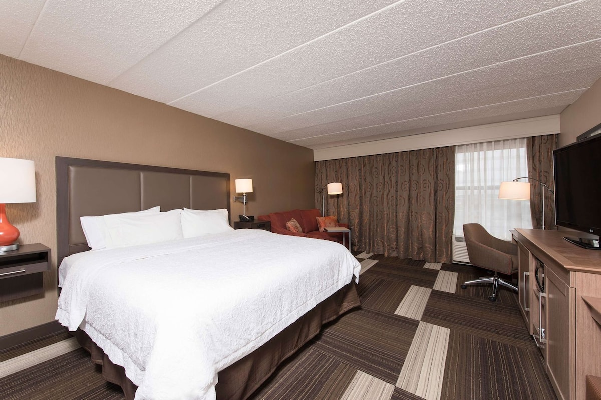 Une chambre d’hôtel avec un grand lit, un bureau, une chaise, une télévision et une fenêtre avec des rideaux.