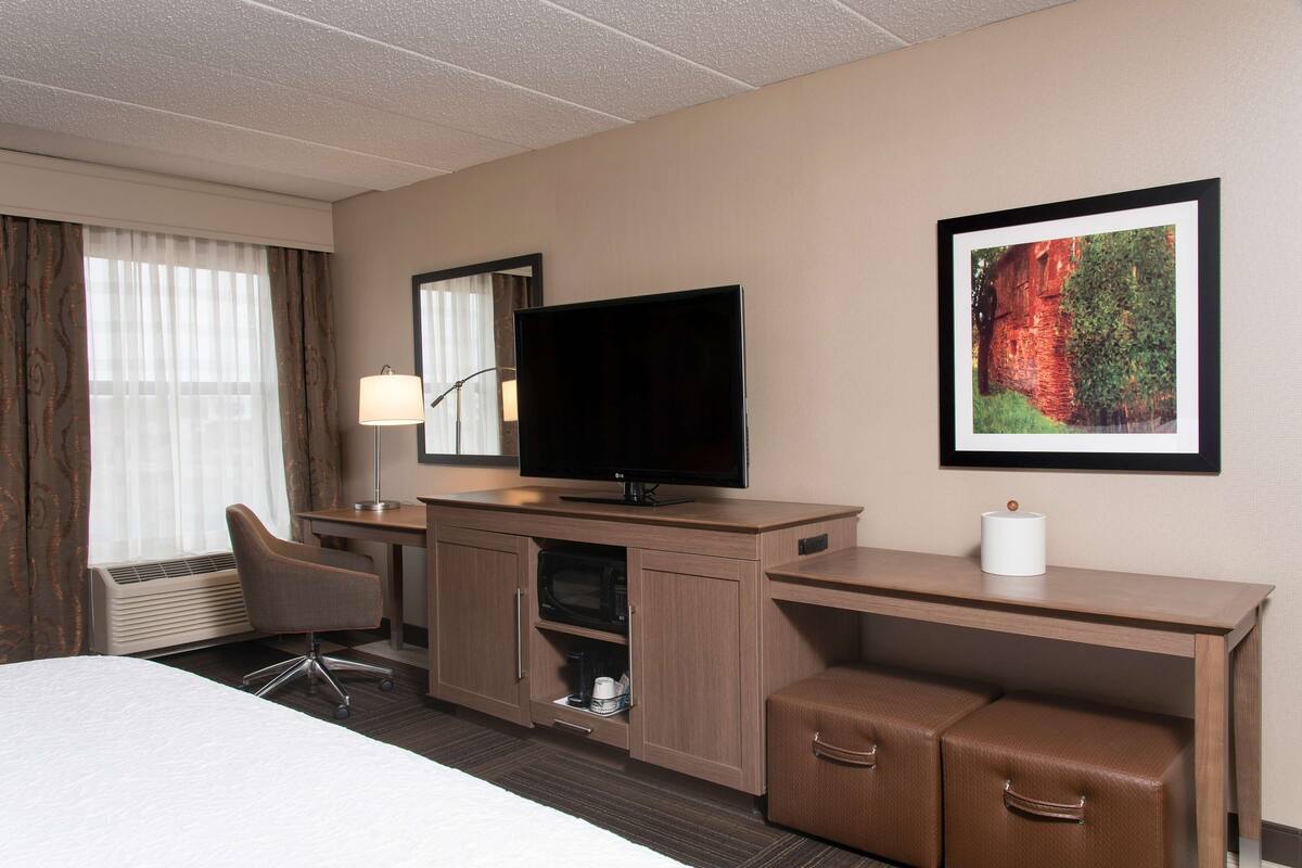 A hotel room with a bed, a desk with a flat-screen TV, a chair, a microwave, and a framed picture on the wall.