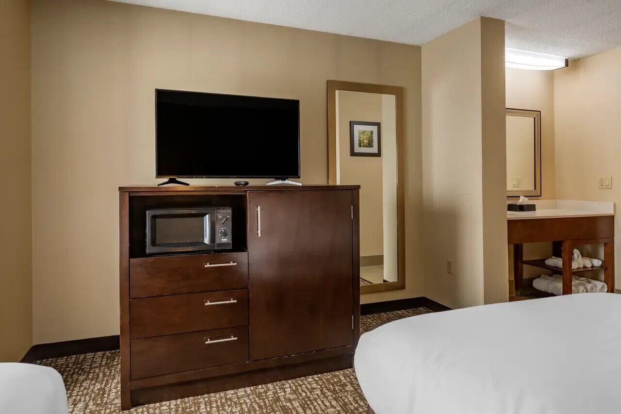 A hotel room with a wooden cabinet, a flat-screen TV, a microwave, a bed, a bathroom with a sink and mirror, and a carpeted floor.