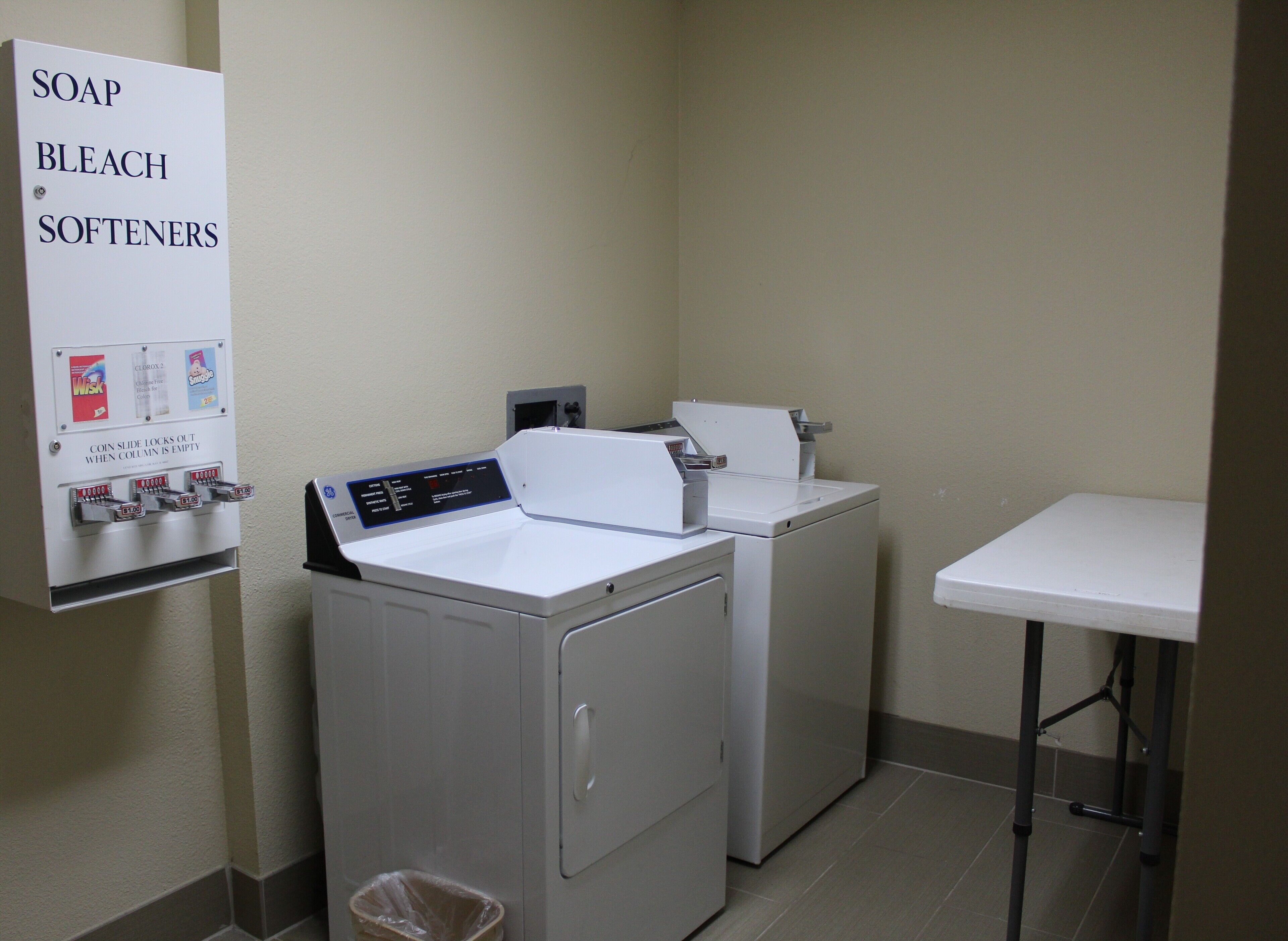 A laundry room with two washers and dryers, a coin-operated soap and bleach dispenser, and a trash bin.
