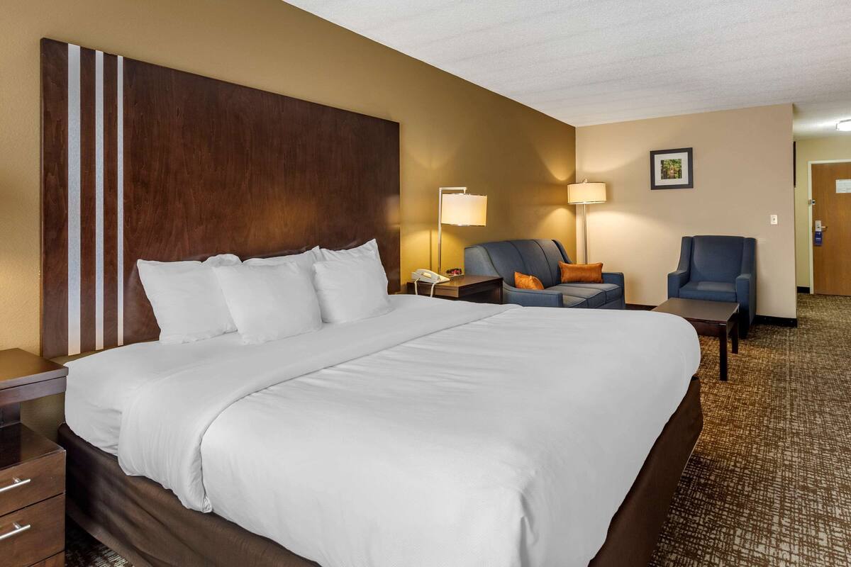 A hotel room with a large bed, a desk with a lamp, two chairs, and a bedside table.