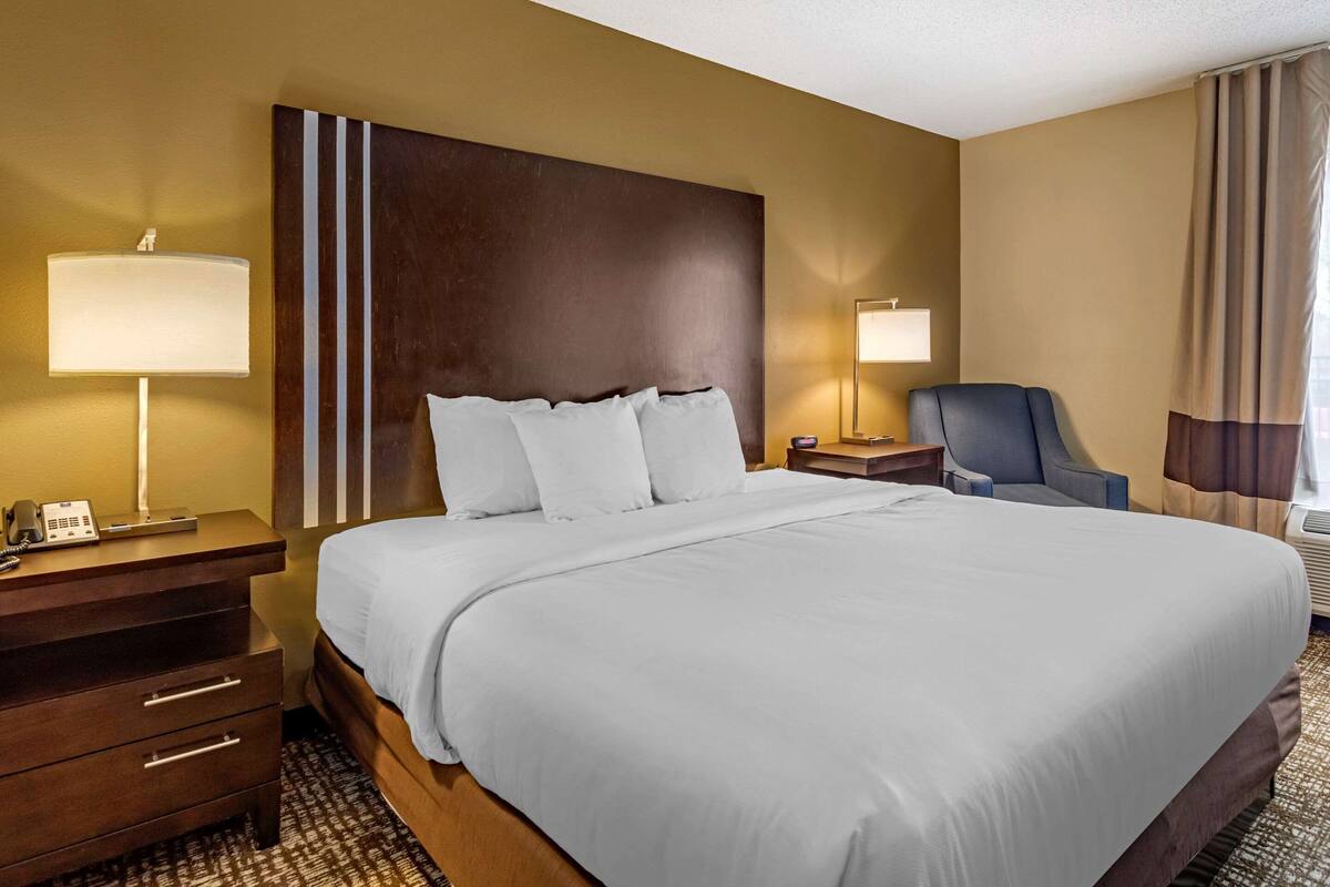 A hotel room with a large bed, a wooden headboard, a desk with a lamp, a chair, and a bedside table.